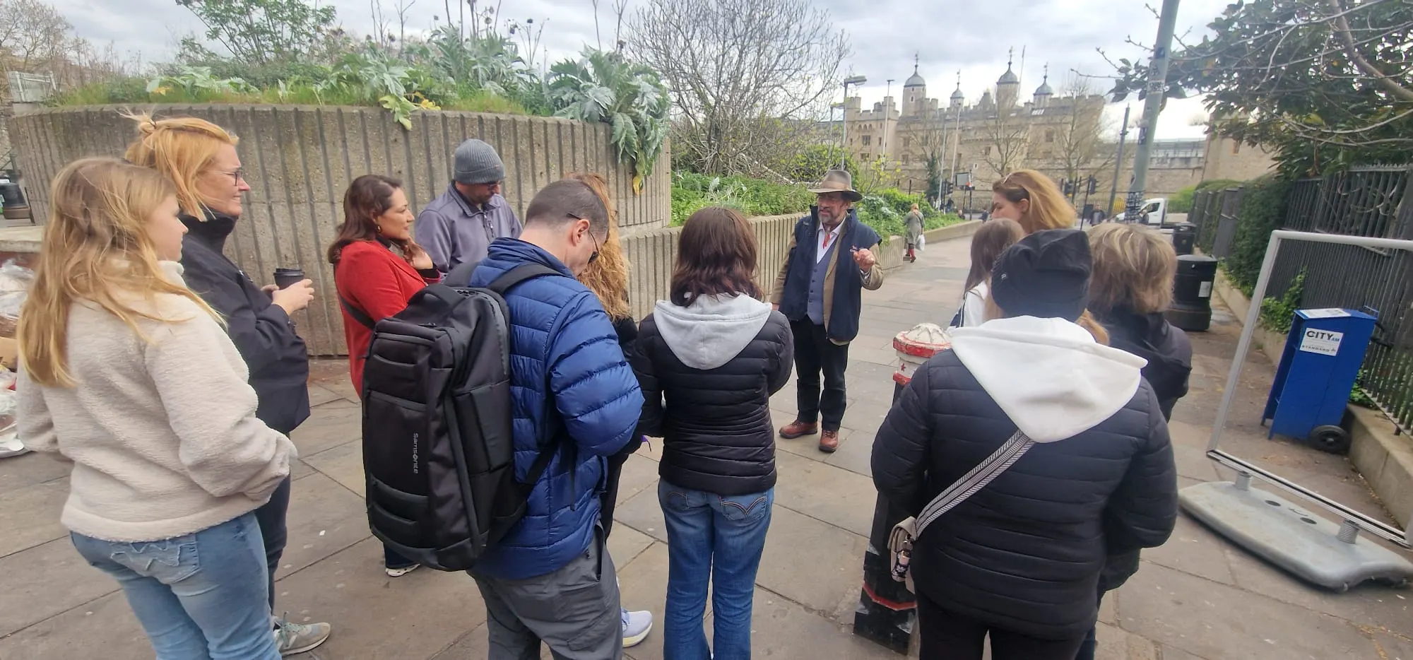 A tour guide in a hat speaks to a group of people with the Tower of London behind them.