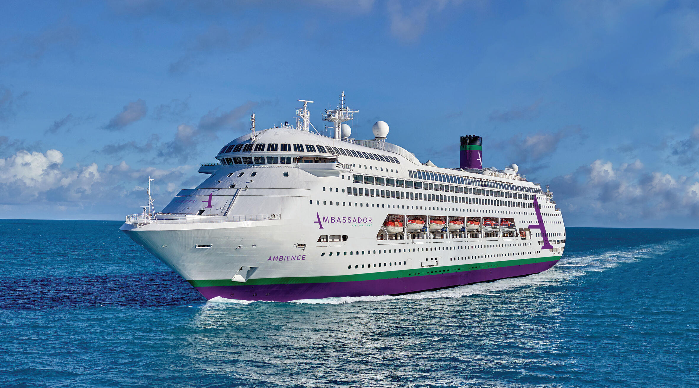 A large white cruise ship with purple and green stripes sailing across a bright blue ocean.