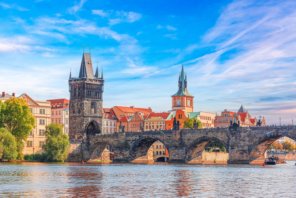 A historic stone bridge with towers crosses a wide river leading to a city under a blue sky.