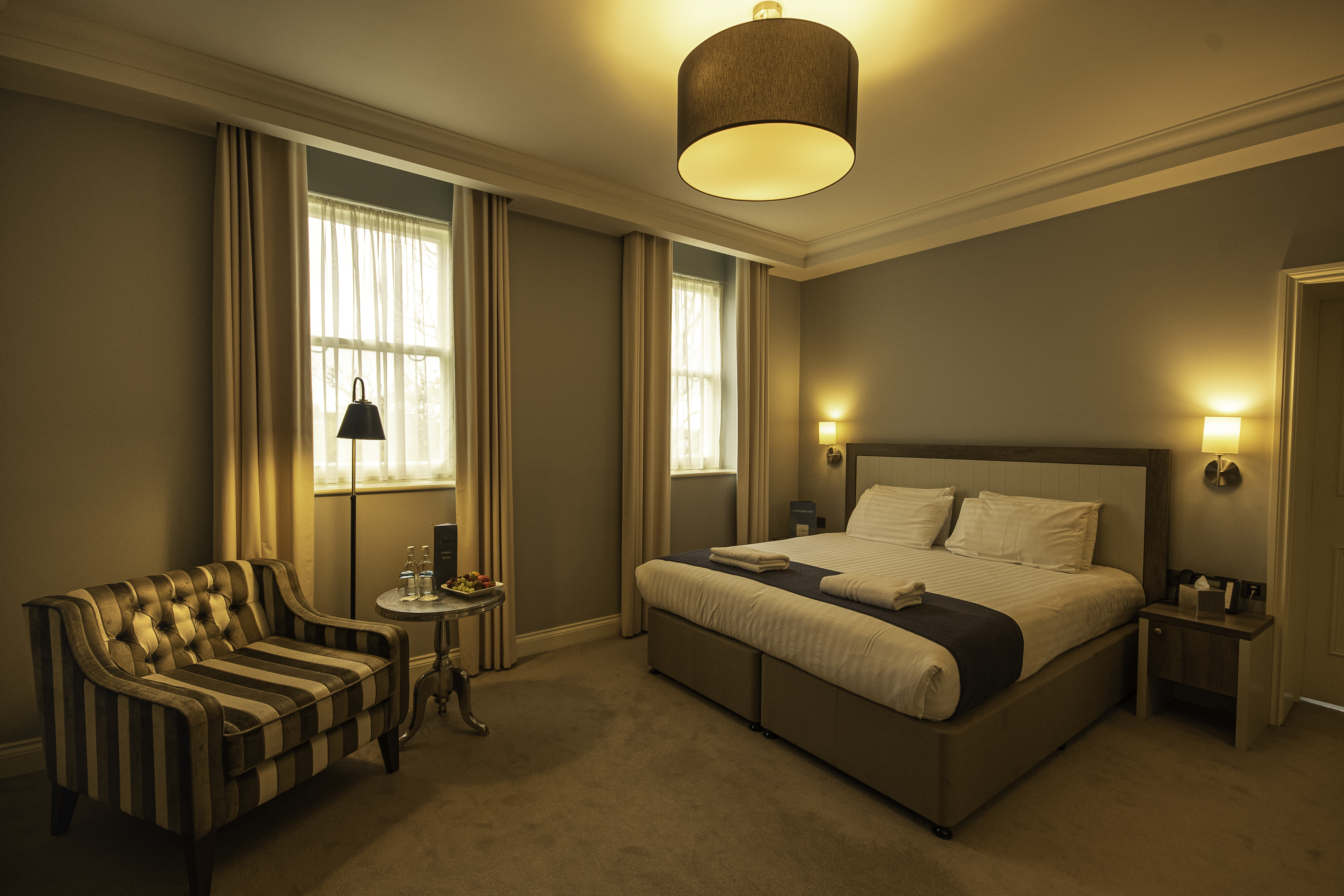 A warmly lit hotel room with a double bed, a striped armchair, and two windows with curtains.