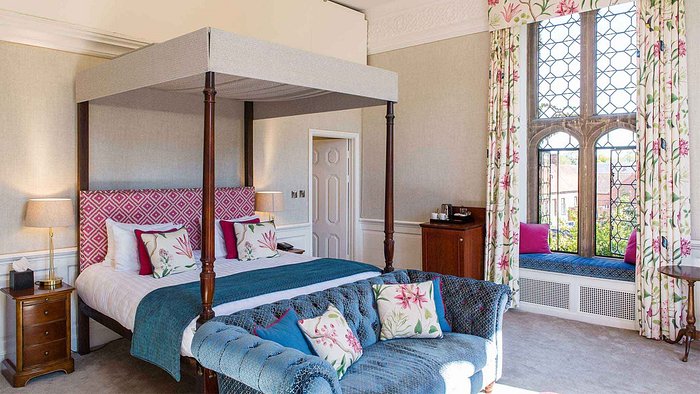 A sunlit hotel room with a four-poster bed, a blue sofa, and a large window with a window seat.