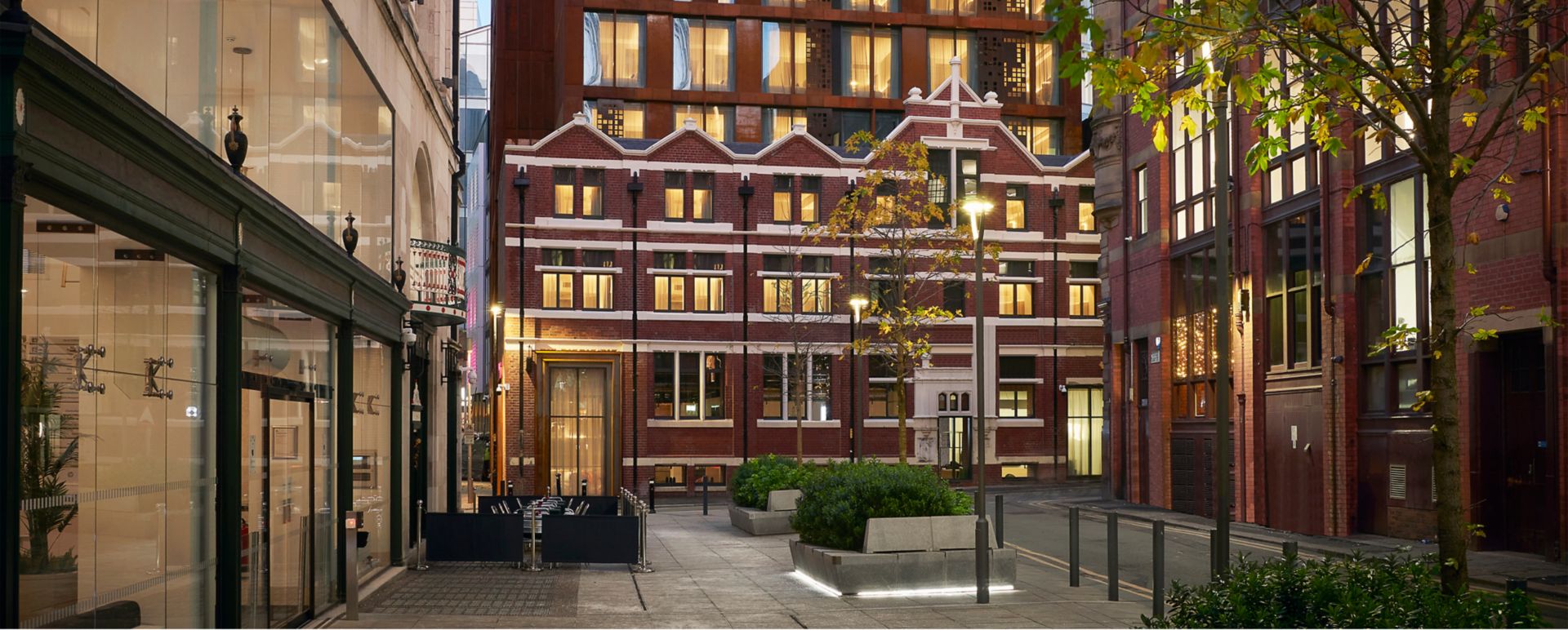 A paved city courtyard at dusk surrounded by illuminated brick and modern buildings with some trees.