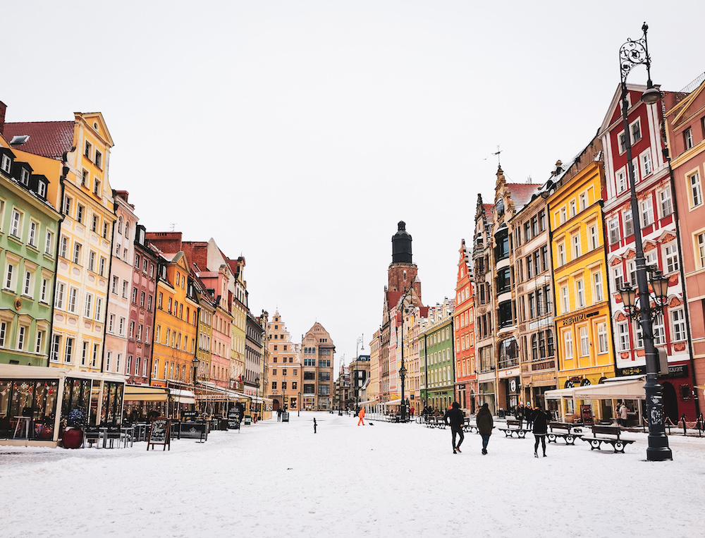 A wide, snow-covered European city square flanked by colorful, multi-story buildings with a few people walking.