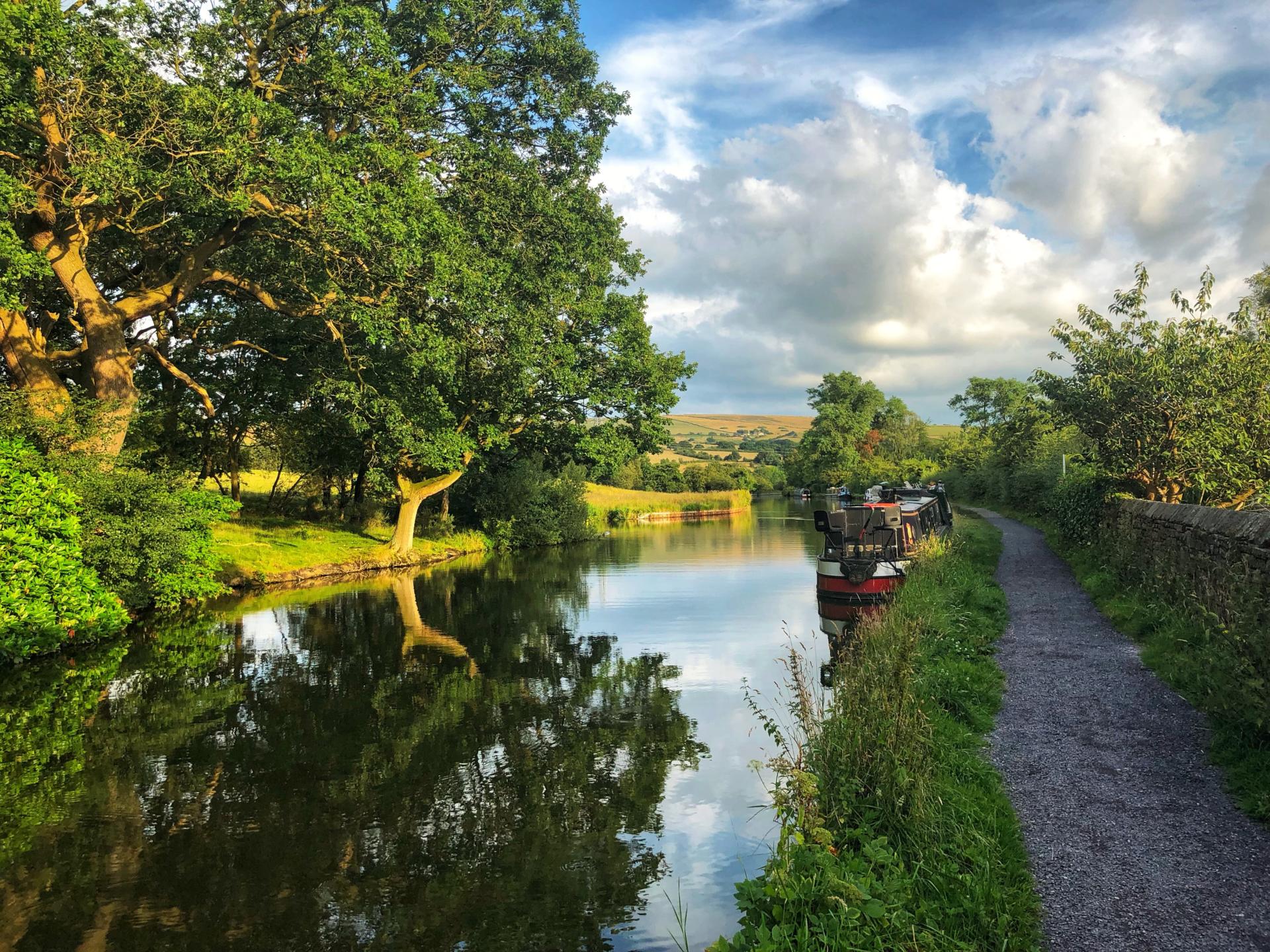 A scenic canal with moored boats, a towpath, and lush trees under a partly cloudy sky.