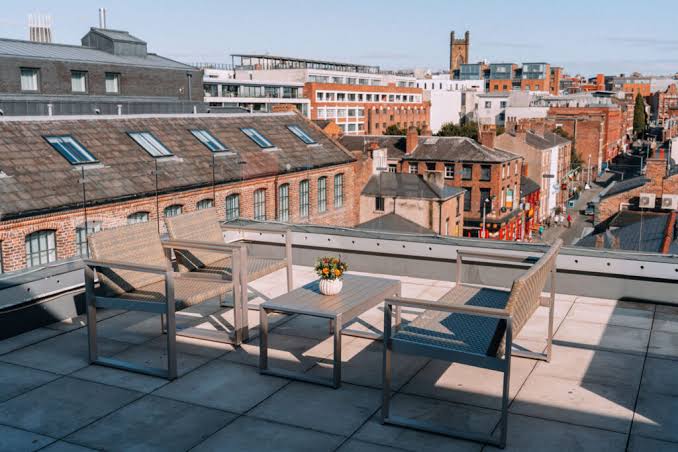 A rooftop patio with chairs and a table, overlooking a city with brick buildings under a clear sky.