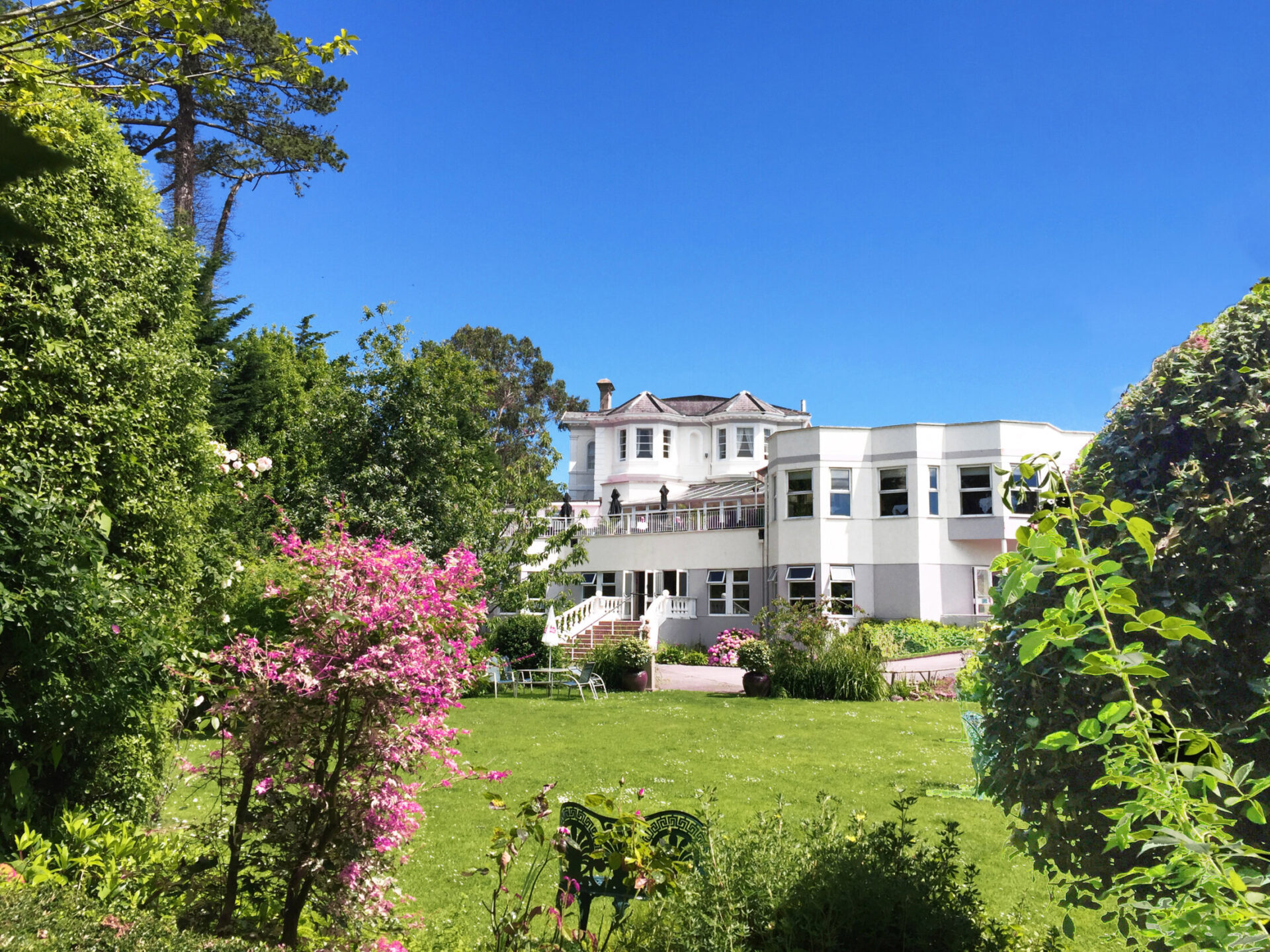 A large white building with a lush green lawn and gardens, seen on a clear, sunny day.