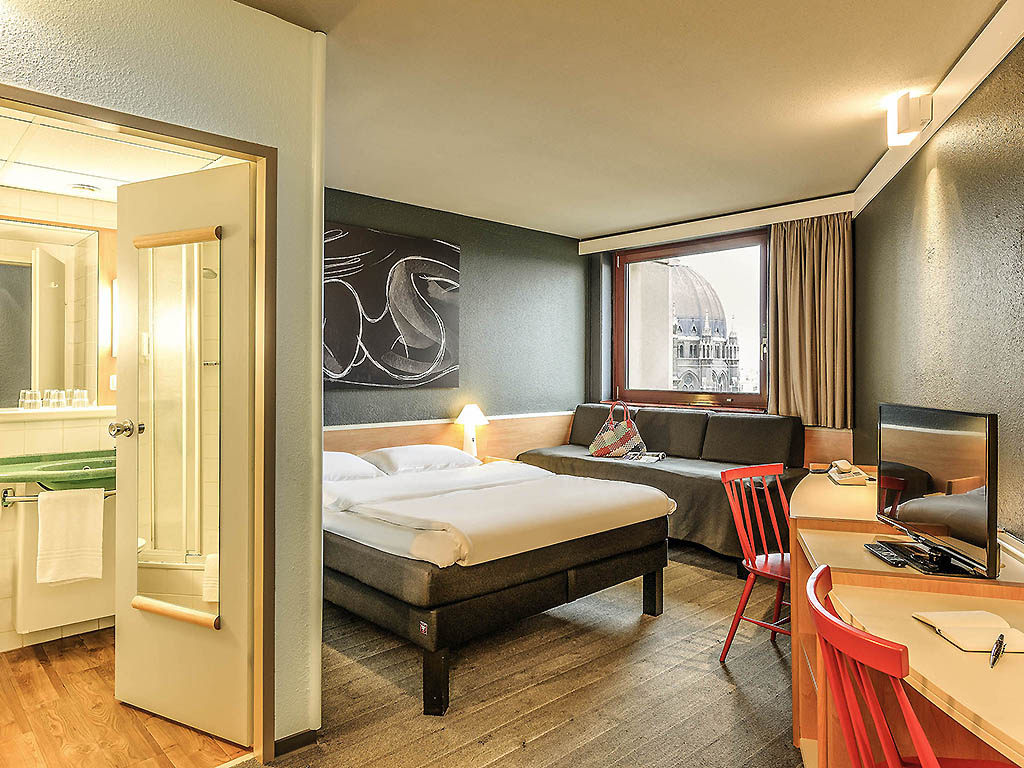 A hotel room with a bed, a desk with a red chair, and a view into the bathroom.