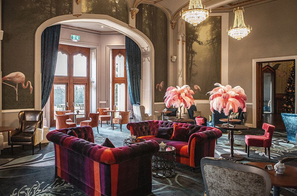 An eclectic, upscale lounge with colorful velvet sofas, feather lamps, chandeliers, and flamingo-themed wall art.