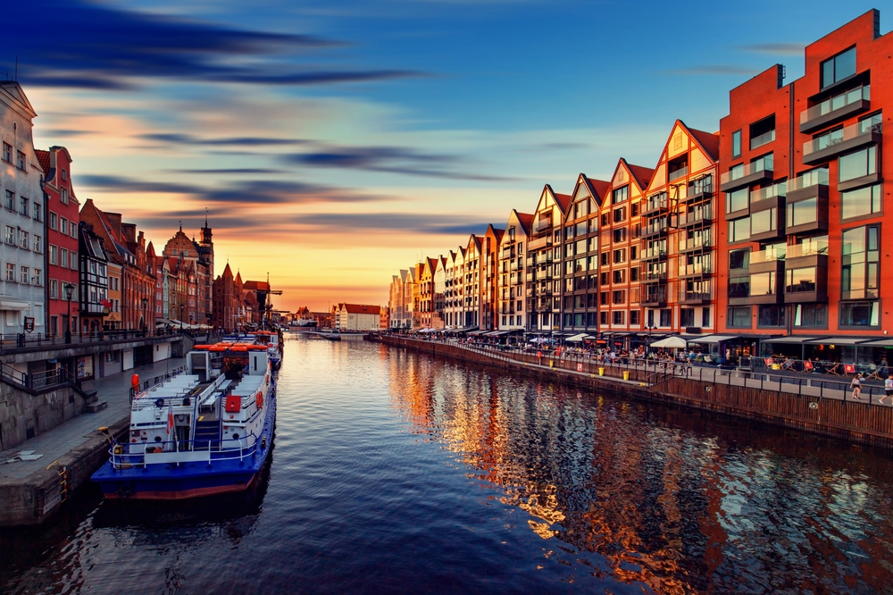 A river runs through a European city with colorful, gabled buildings lining the banks at sunset.