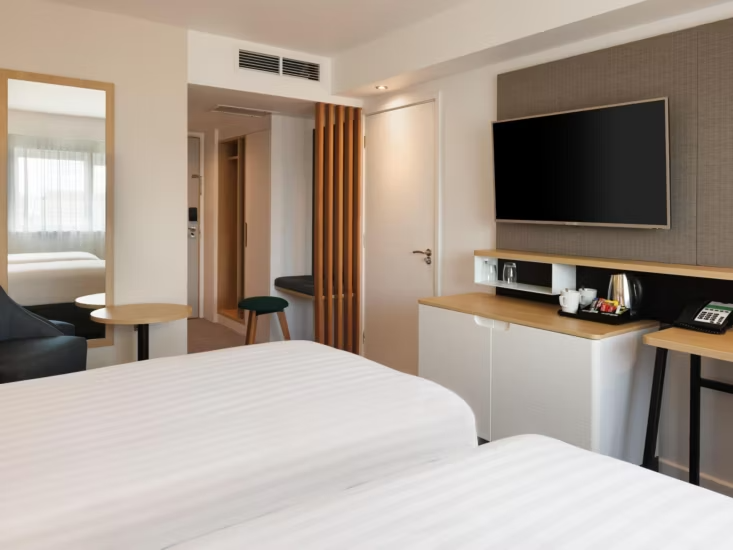 A modern hotel room with two twin beds, a wall-mounted television, and a desk area.