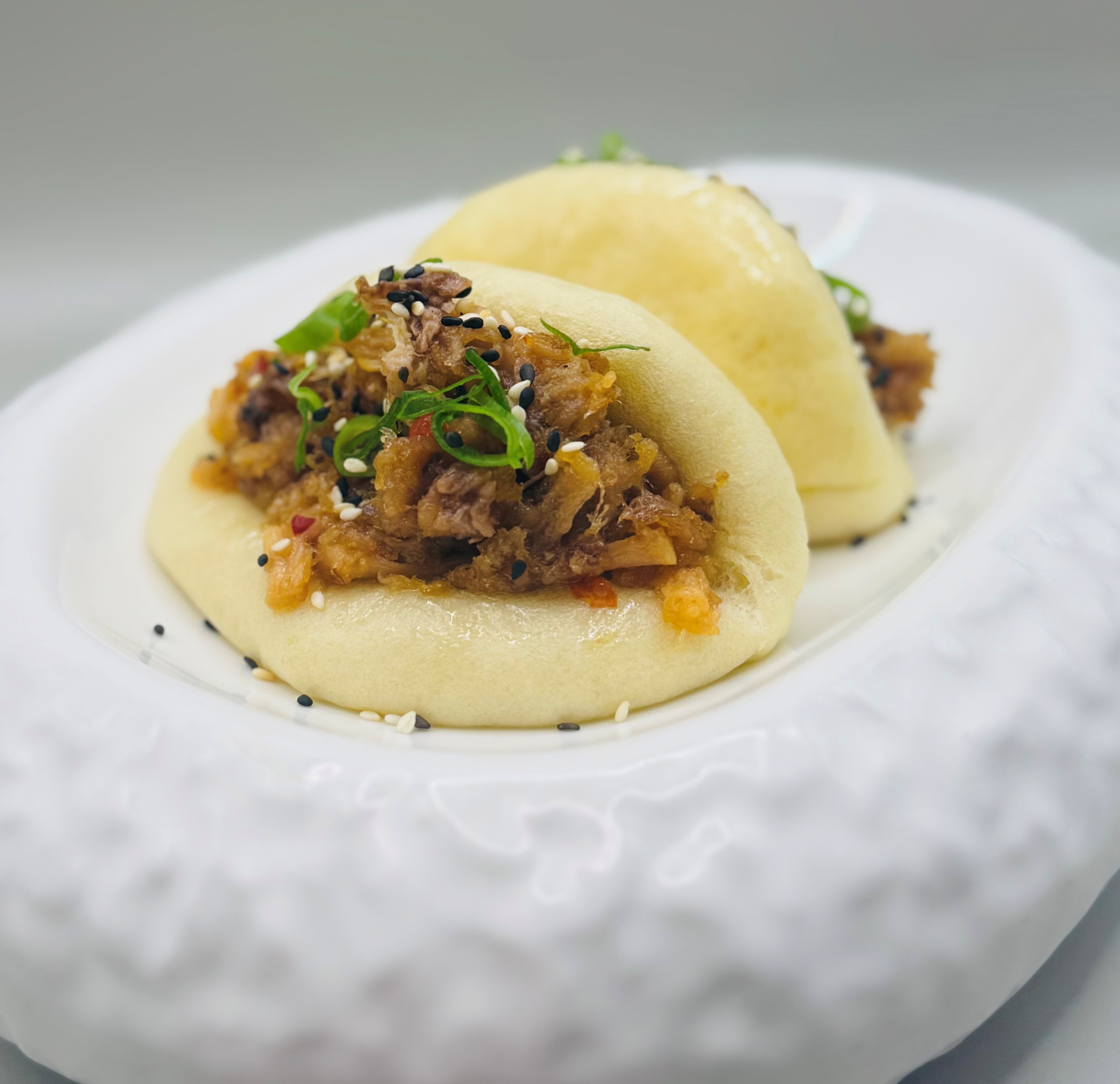 Two steamed bao buns with shredded meat filling, garnished with scallions and sesame seeds on a white plate.