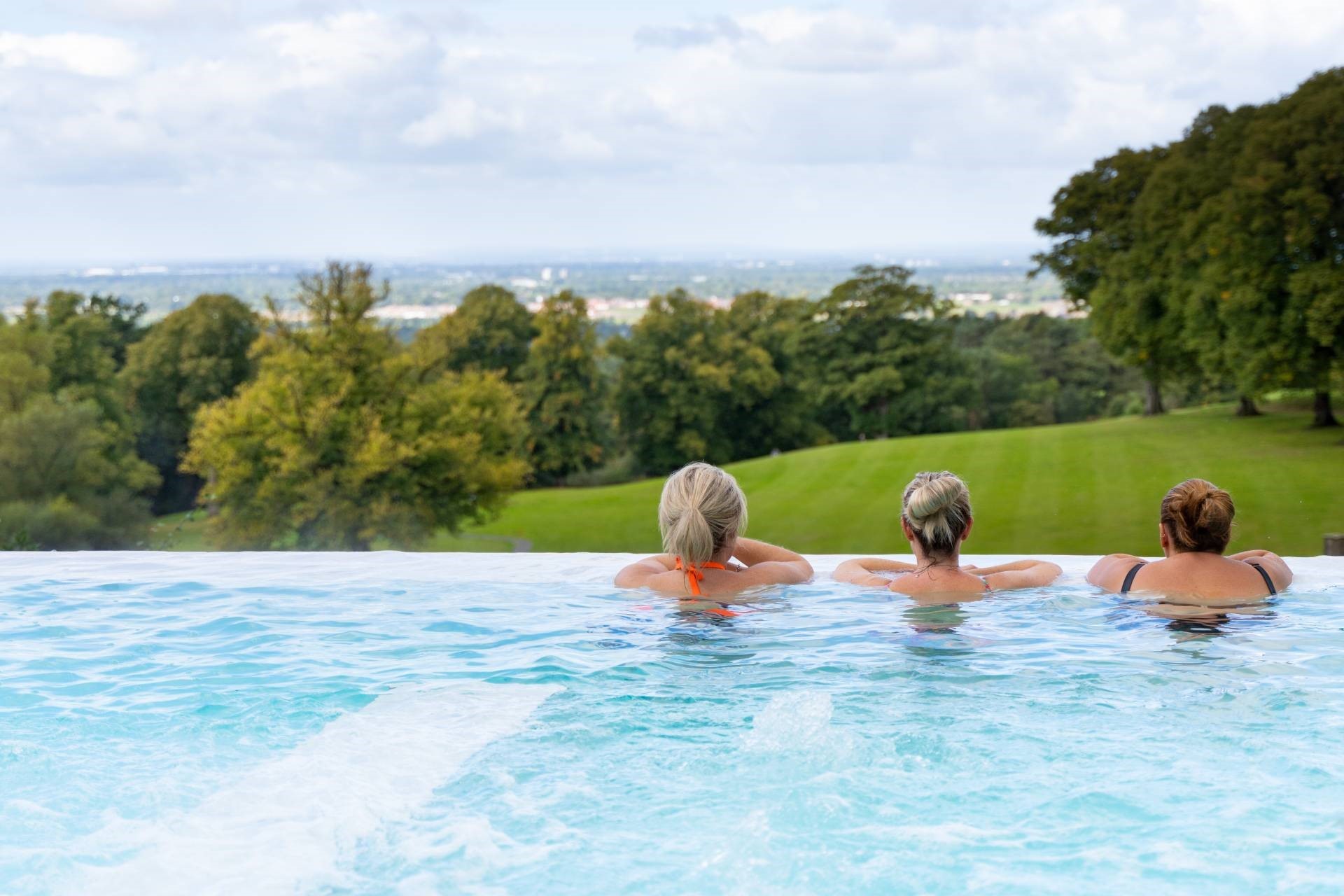 Three women seen from behind relaxing in an infinity pool overlooking a green, tree-filled landscape.