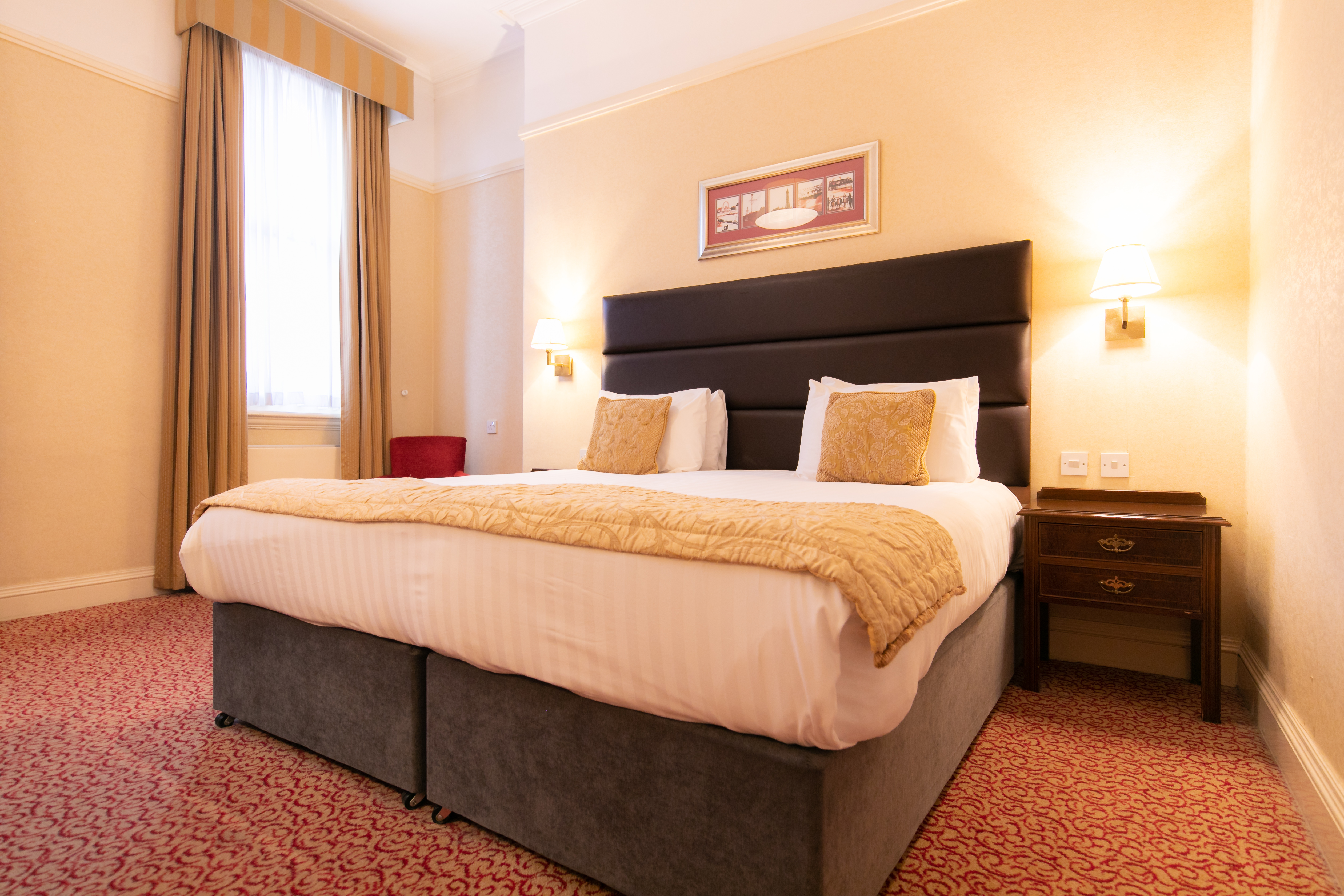 A neatly made double bed in a hotel room with a large window and patterned red carpet.