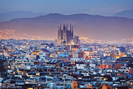 The Barcelona skyline at dusk, with the Sagrada Família cathedral lit up and mountains in the background.