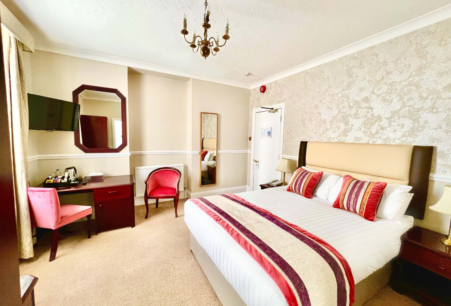 A hotel room with a double bed, a desk, a television, and two pink chairs.