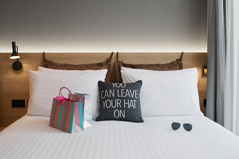 A made bed with shopping bags, sunglasses, and a decorative pillow saying 'YOU CAN LEAVE YOUR HAT ON'.