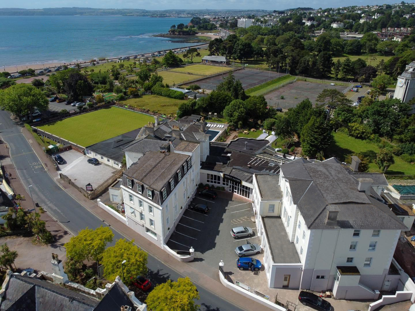 Aerial view of a coastal hotel complex with lawns, tennis courts, and the sea in the background.