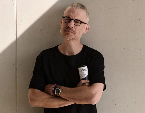 Man with glasses and a beard, wearing a black shirt, holding a can, standing against a wall.