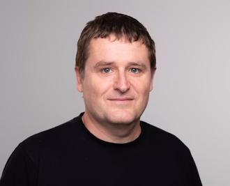 A person with short hair wearing a black shirt against a gray background.