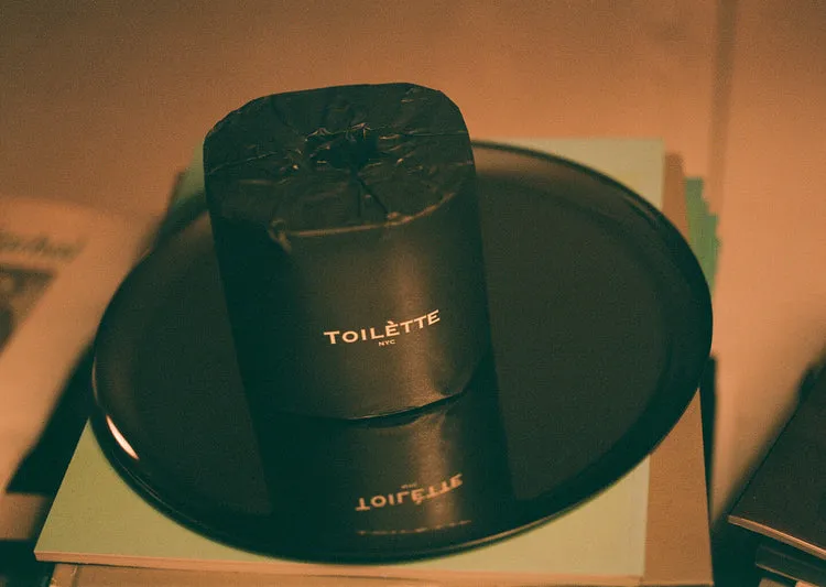 A roll of toilet paper in a black wrapper that reads 'TOILÈTTE NYC' sits on a black plate.