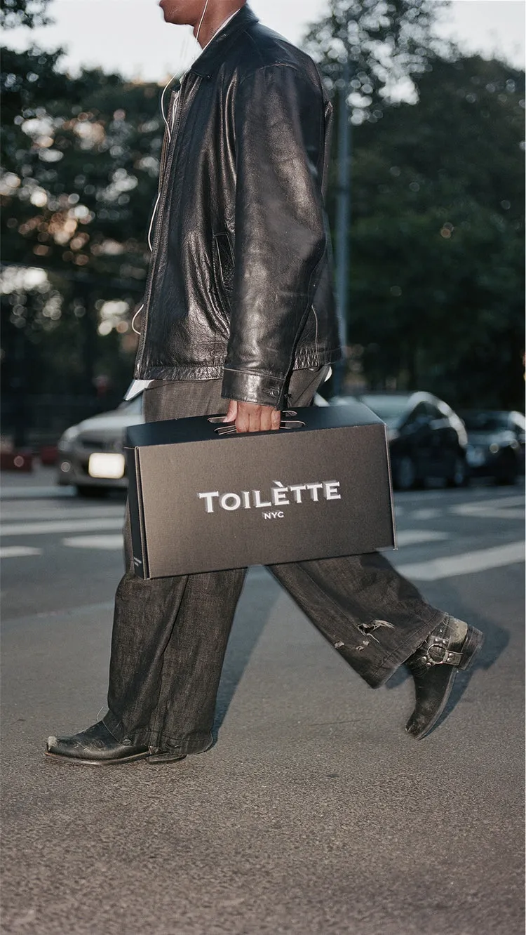 A person in a black leather jacket and jeans walks across a street carrying a box.