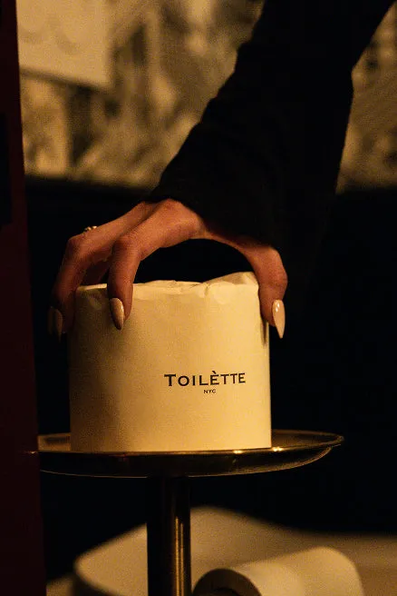 A hand with long manicured nails places a paper-wrapped toilet roll labeled 'TOILÈTTE NYC' on a gold stand.