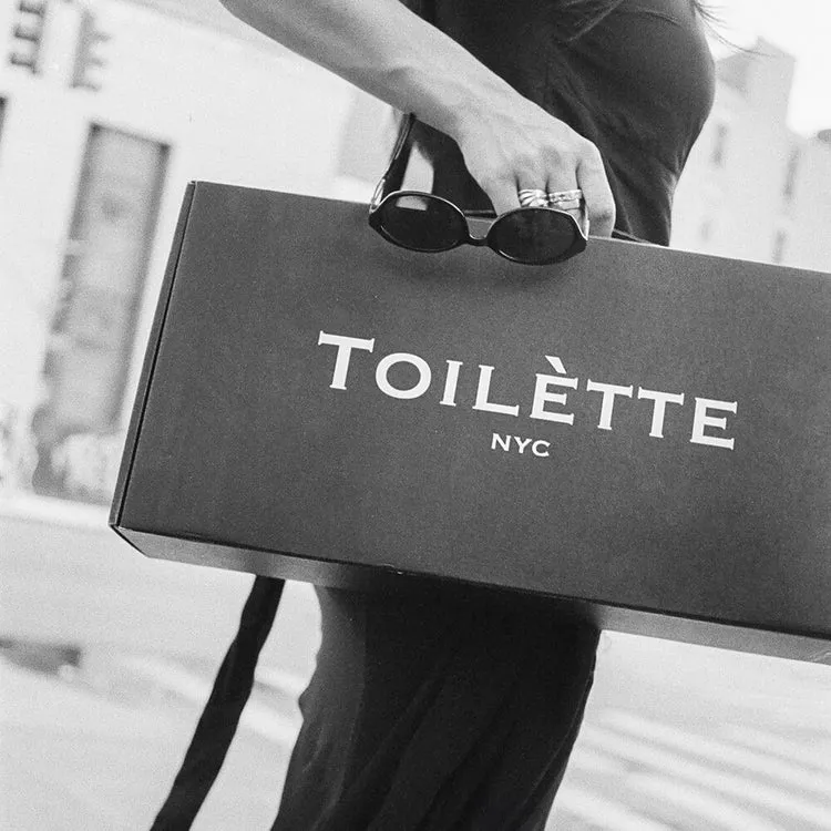 A person in black and white holding a large black box that reads 'TOILÈTTE NYC' and sunglasses.