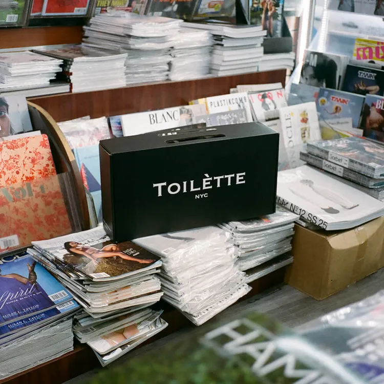 A black box with the words 'TOILÈTTE NYC' sits on a pile of magazines in a newsstand.