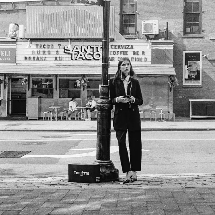 A black and white photo of a woman in a suit standing on a city street in front of a restaurant.