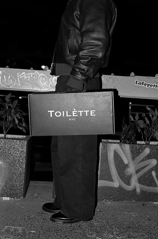 A person in a black leather jacket at night holds a black box that reads 'TOILÈTTE NYC'.