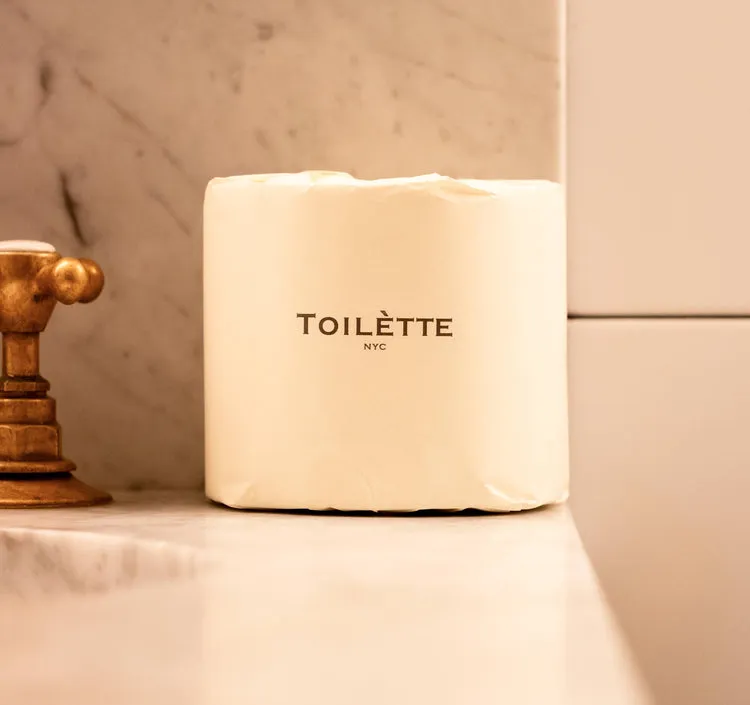 A wrapped roll of Toilètte NYC toilet paper sitting on a marble countertop next to a faucet.