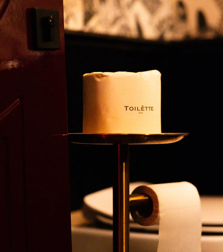 A brass toilet paper stand holding a wrapped roll with 'TOILÈTTE NYC' printed on the side.
