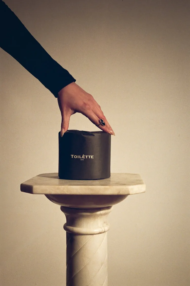 A hand rests on a roll of black toilet paper sitting on a marble pedestal.