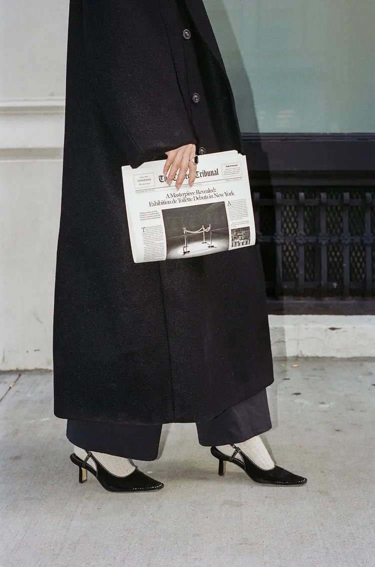 A person in a long black coat and black heels holds a folded newspaper on a sidewalk.