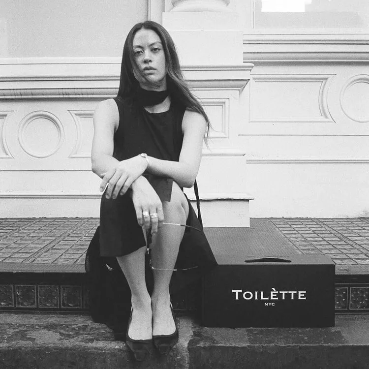 A black and white photo of a woman in a black dress sitting on steps next to a box.