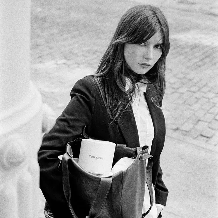 A black and white photo of a woman holding a tote bag with a roll of toilet paper in it.