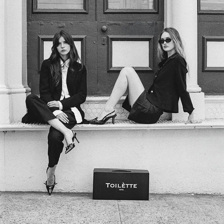 A black and white photo of two women in black outfits sitting on a ledge with a shoebox.