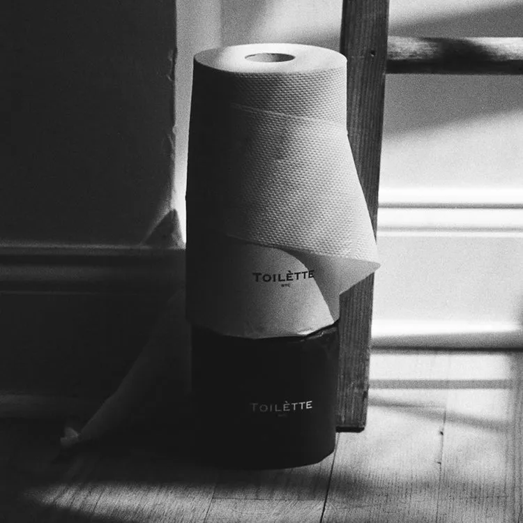 A black and white photo of a roll of toilet paper branded 'TOILETTE' sitting on a container.