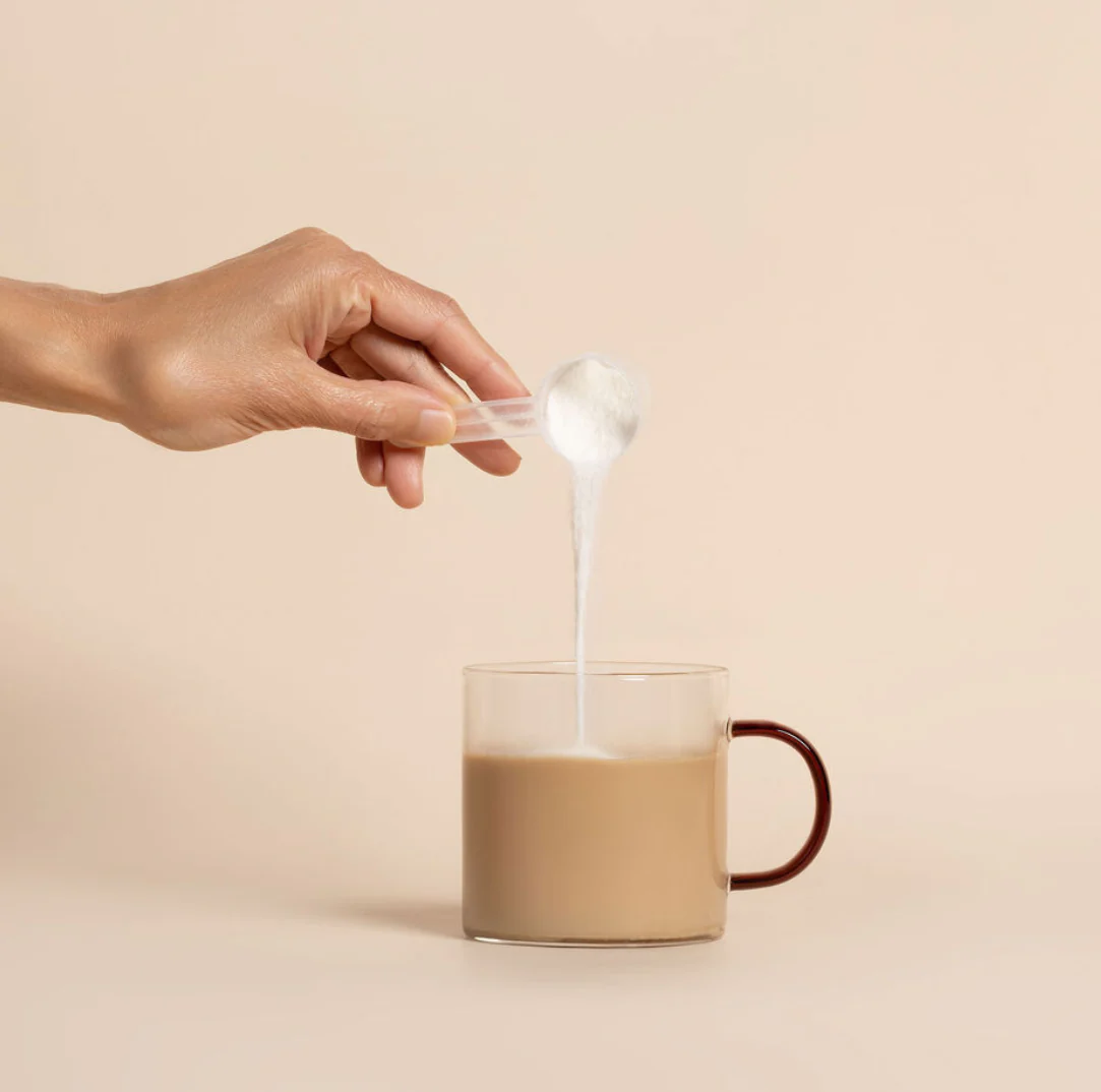 Hand pouring powdered creamer into a beige cup of coffee against a neutral background.