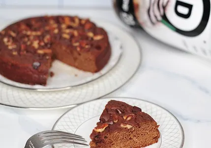 A slice of chocolate cake topped with nuts on a plate, with the rest of the cake behind.