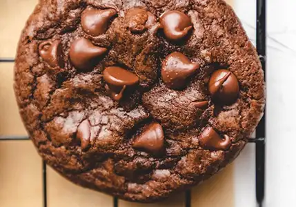 A close-up of a double chocolate chip cookie resting on a black wire cooling rack.