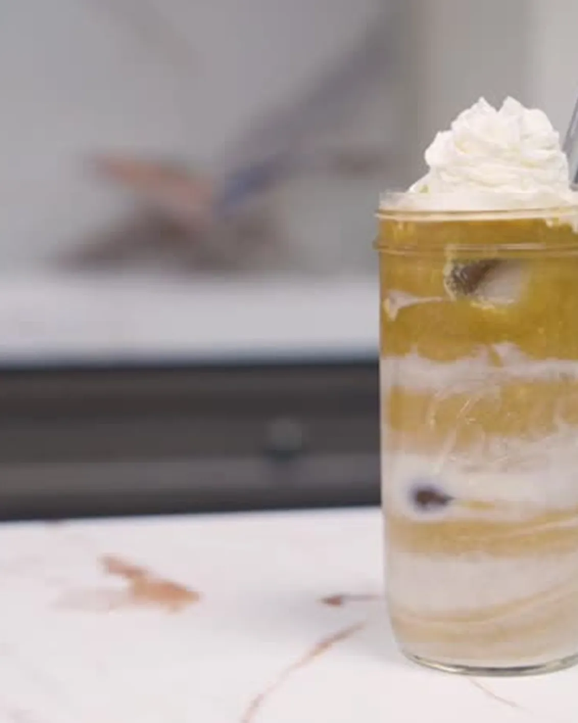 A close-up of a layered drink in a glass jar, topped with whipped cream on a marble countertop.