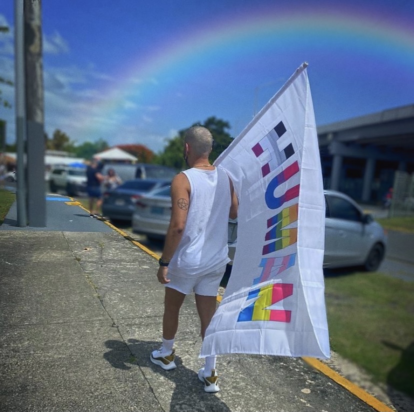 Person in white holding a 'HUMAN' flag with rainbow colors, under a rainbow.