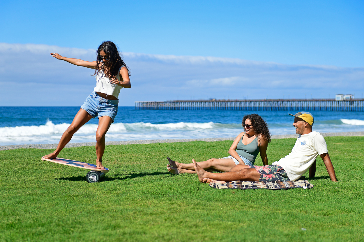 A woman uses a balance board on the grass while two friends watch from a blanket by the sea.