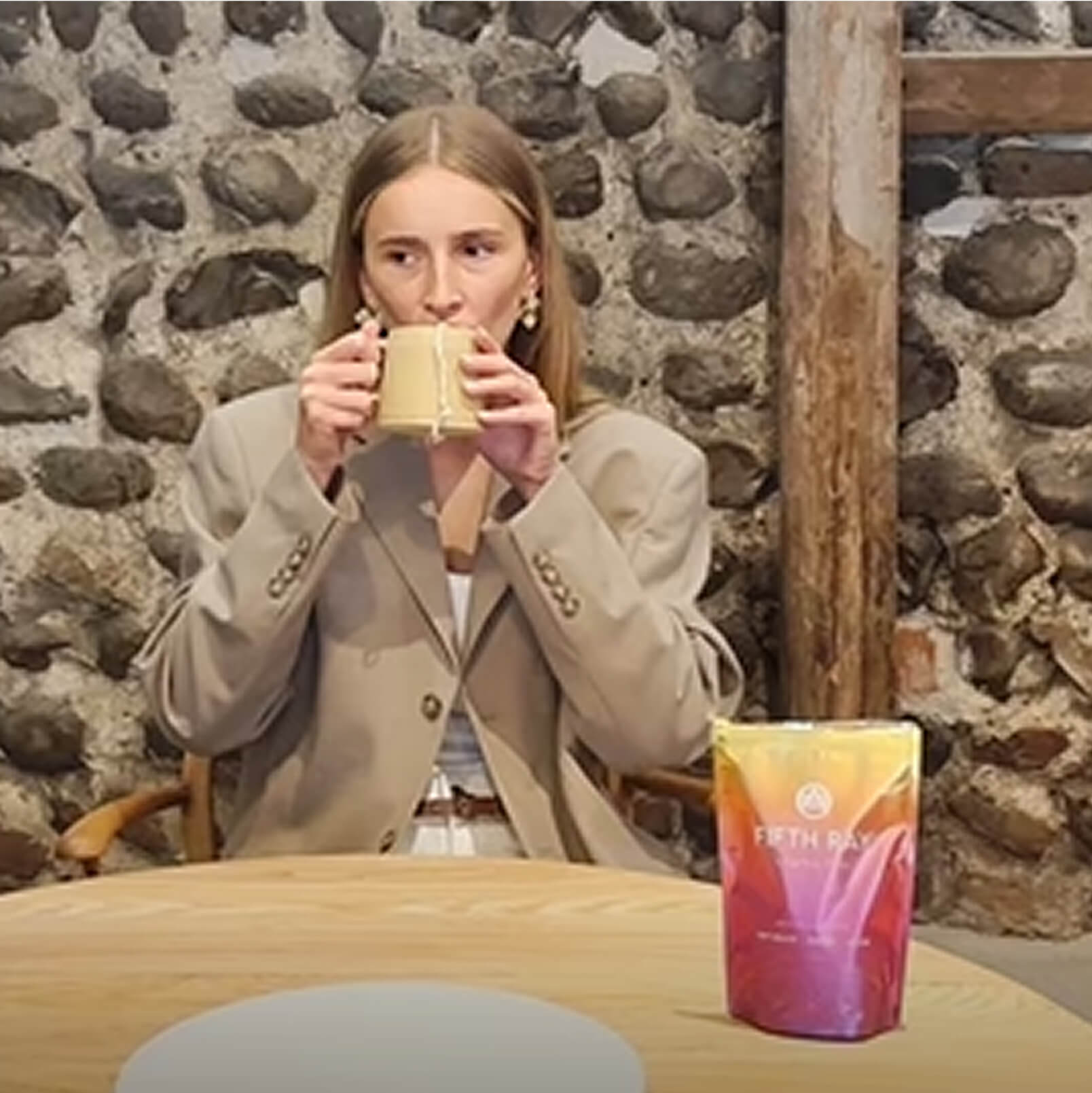 Person in a beige blazer drinking from a mug, with a colorful package on the table.