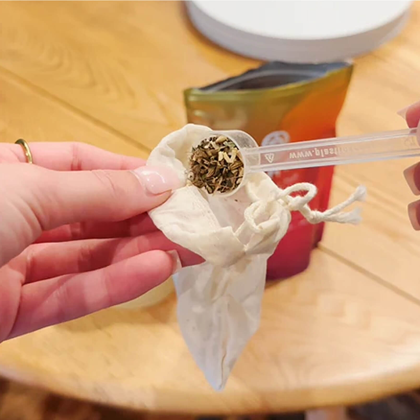 Hand holding a scoop of herbs over a cloth bag on a wooden table.