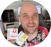A smiling bald man in a floral shirt holding up two small packets.