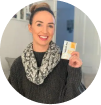 A smiling woman wearing a sweater and scarf holds up a small, white and yellow packet.
