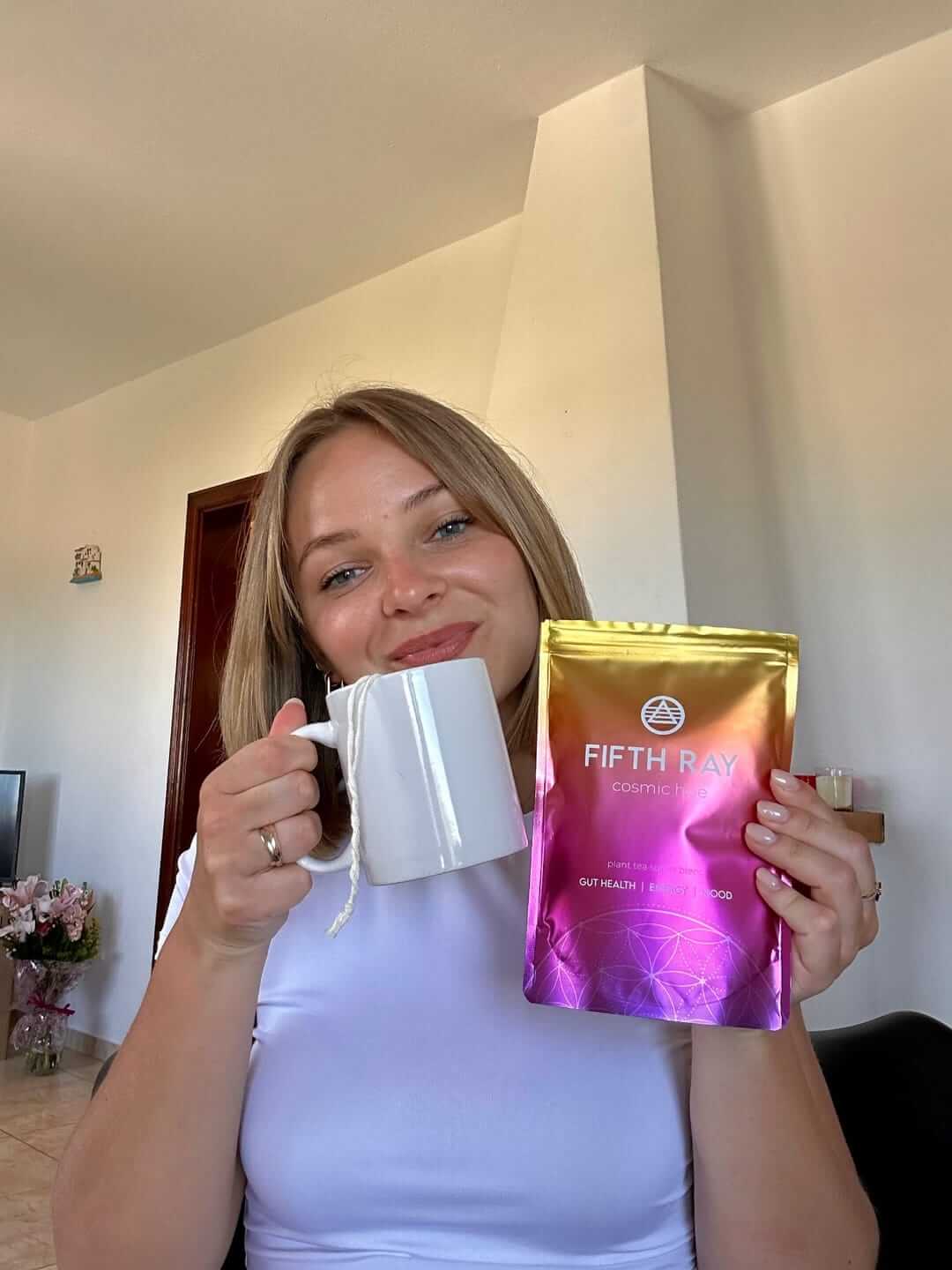 Person holding a white mug and a colorful tea package labeled 'Fifth Ray Cosmic Tea.'