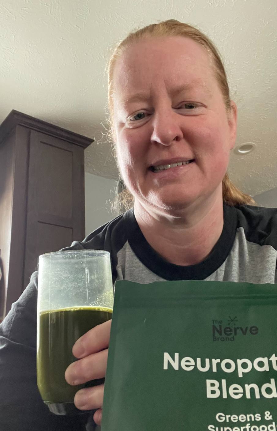A woman smiles while holding a glass of green liquid and a pouch of Neuropathy Blend.